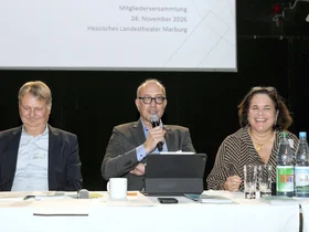 Der Vorsitzer Apostolos Tsalastras und die Geschäftsführung des LMV, Sebastian Kuth und Monika Menezes-Kuth auf dem Podium bei der Mitgliederversammlung 2025.