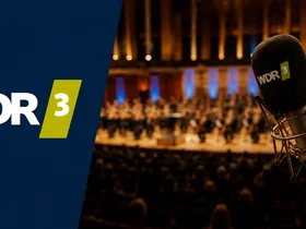Auf der linken Seite: das WDR3-Logo.
Auf der rechten Seite: ein Mikrofon mit WDR3-Branding im Vordergrund; im Hintergrund verschwommen zu sehen ein Orchester auf einer Bühne in einem voll besetzten Konzertsaal.